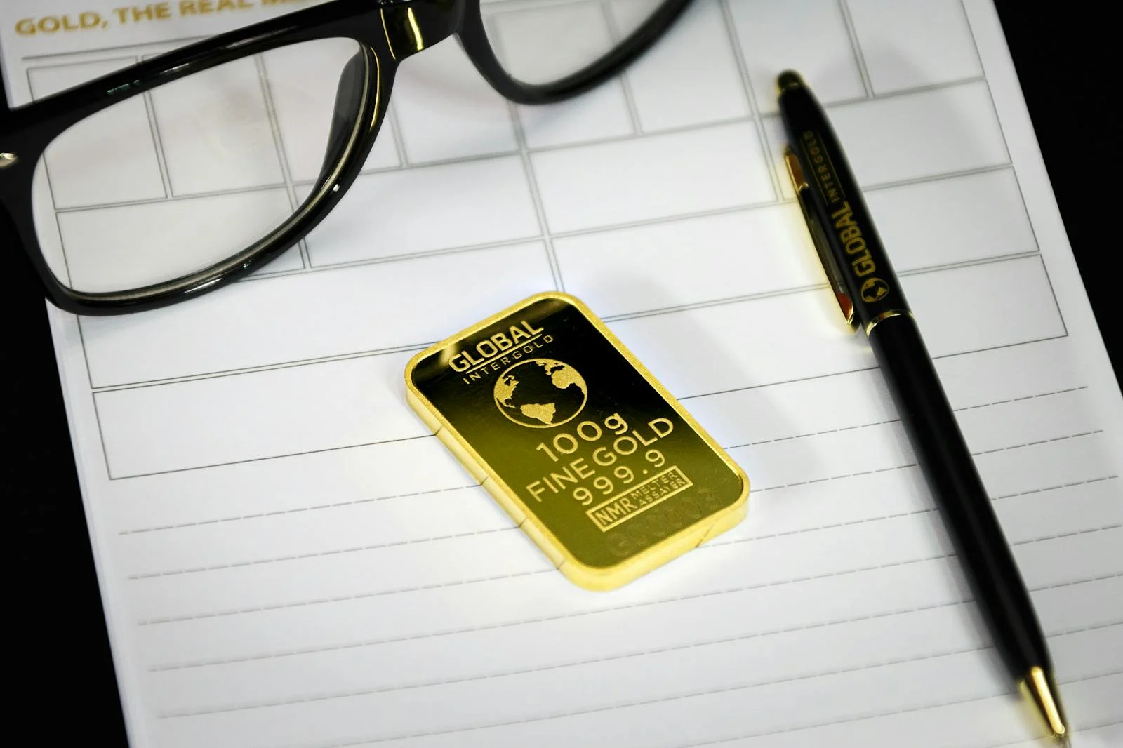 Photo by Michael Steinberg Gold bullion on notepad with eyeglasses and pen, symbolizing wealth and business planning.