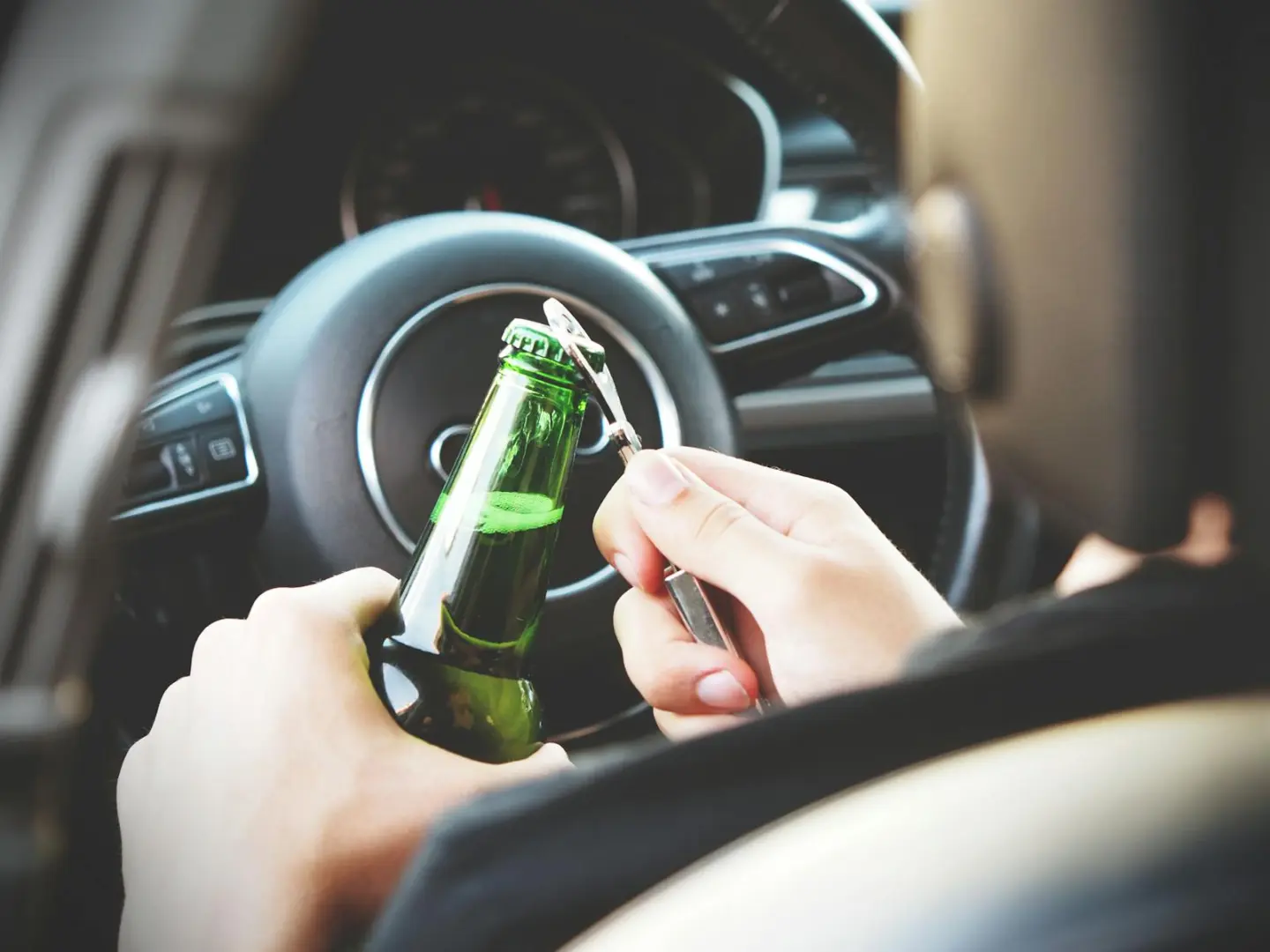 Hands opening a beer bottle inside a car, highlighting the importance of safe driving and awareness of road dangers.